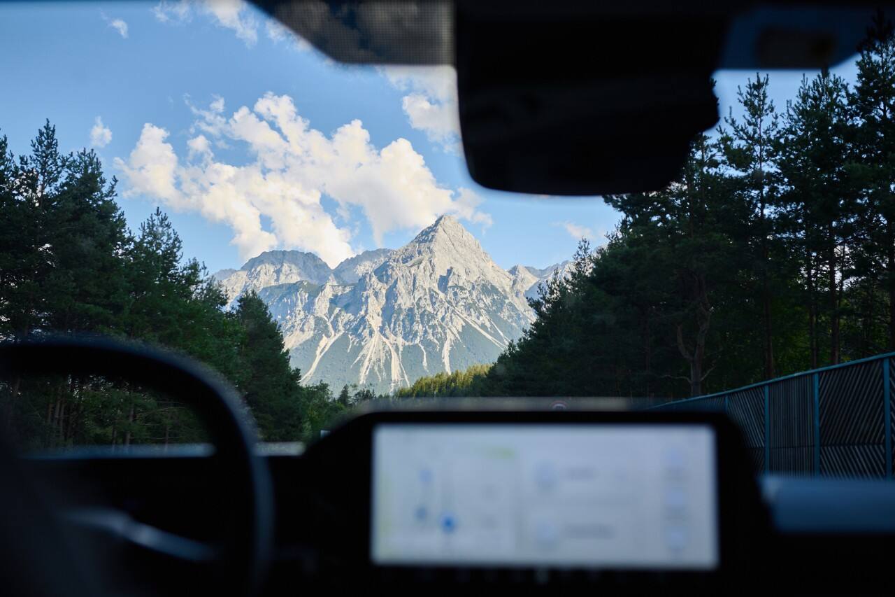 A view through a windshield from inside a vehicle of a mountain with a blue sky and white clouds above.